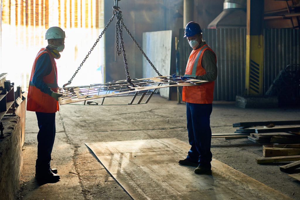 Twee productiemedewerkers met veiligheidsuitrusting begeleiden een stalen constructie aan hijskettingen in de fabriek tijdens het verzinken van staal